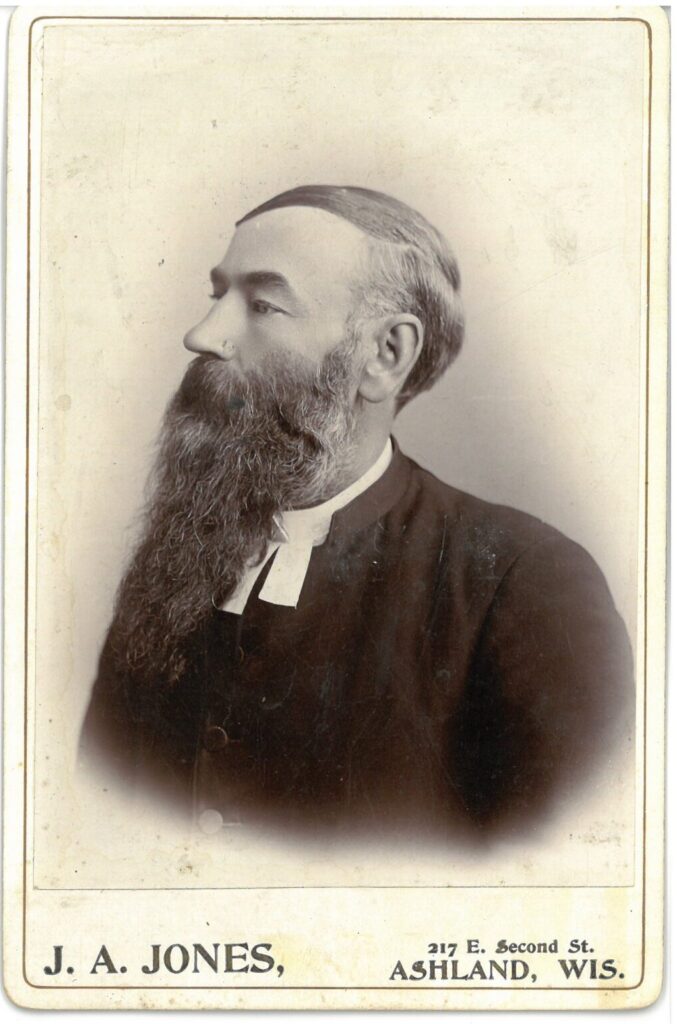 Sepia colored photo of a young man with very long beard wearing pastor's collar.