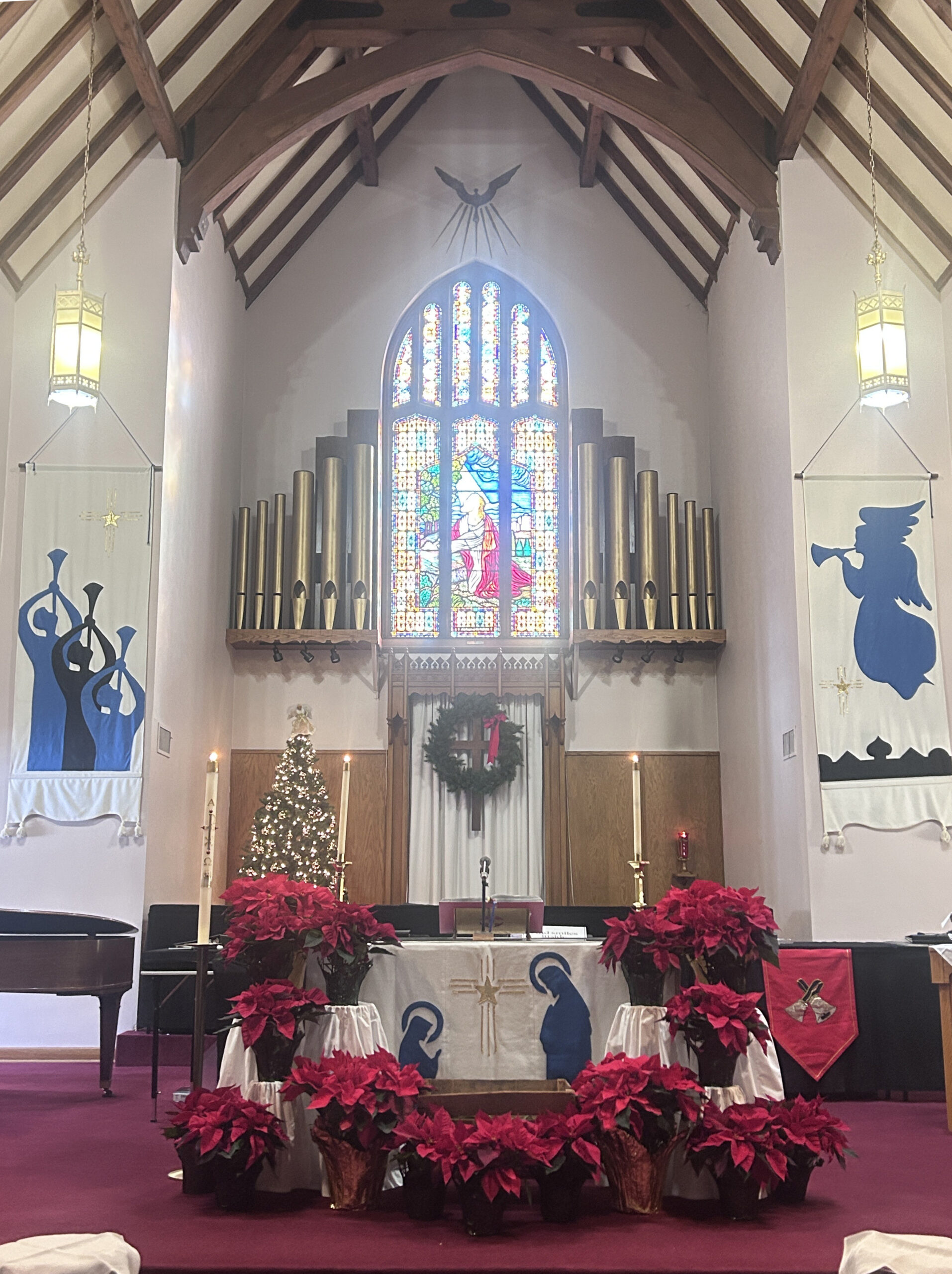 Festive Christmas Altar with angel banner, trumpet banner, altar cloth with kneeling Mary and Joseph, and poinsettias in front.