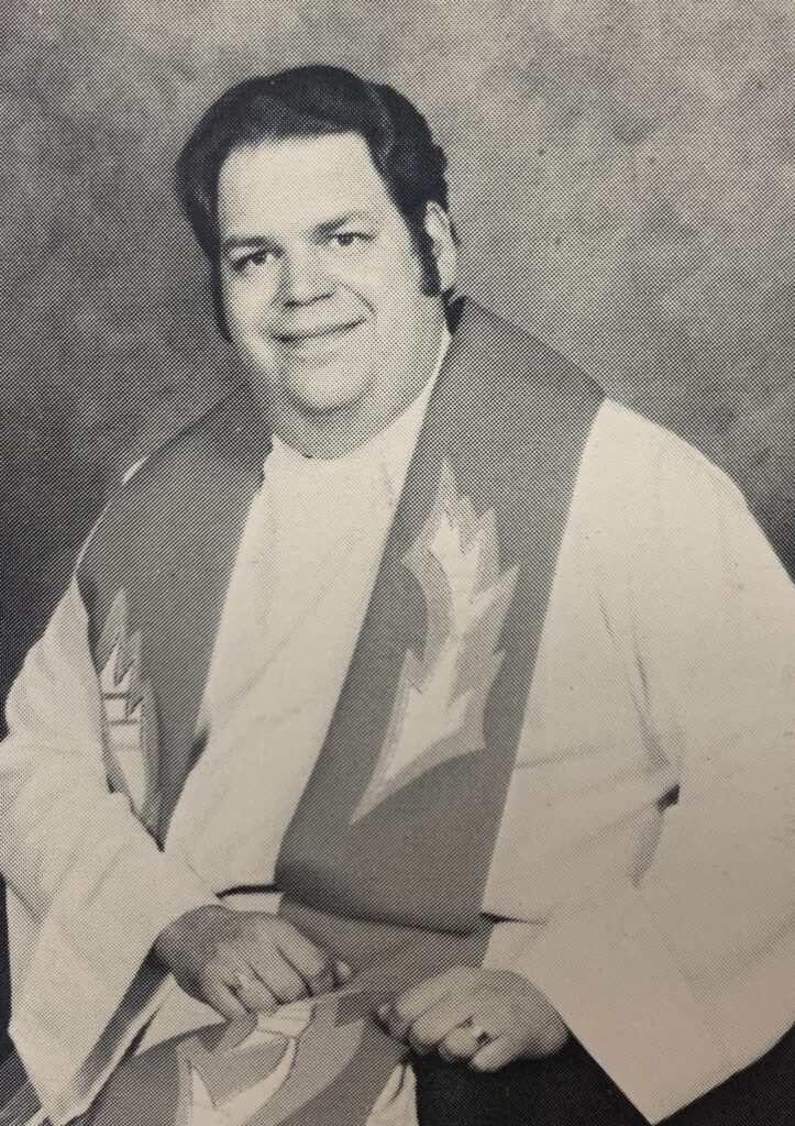 Large man with dark hair and sideburns wearing white clerical robes and stole with fire embroidered on it.