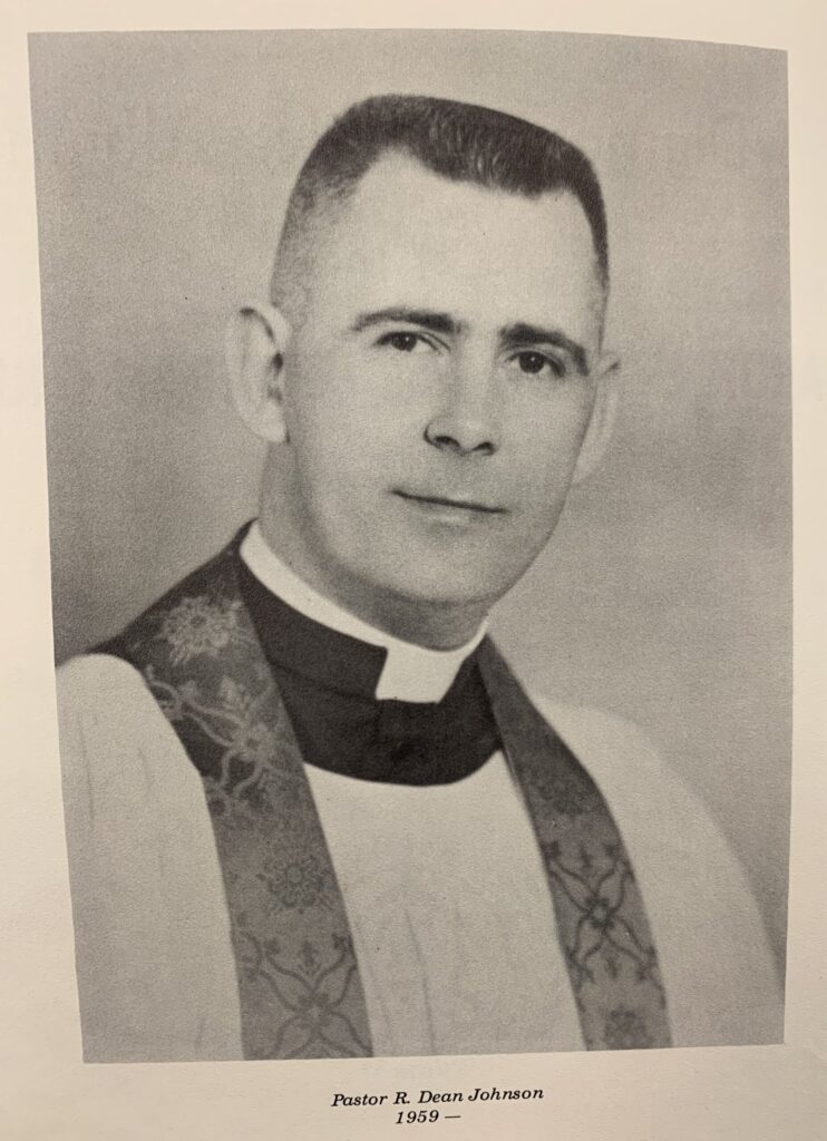 Black and white portrait of man with buzz cut, white robe, clerical collar, and church garments.