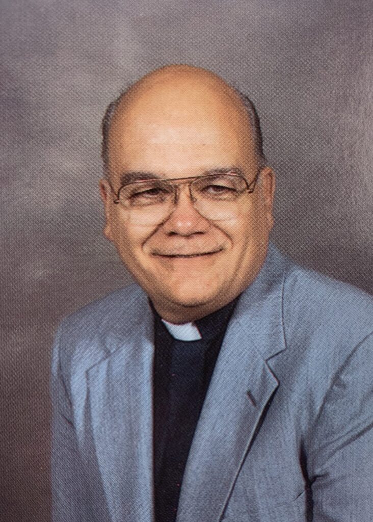Color portrait of balding man wearing black shirt, clerical collar and light blue jacket.
