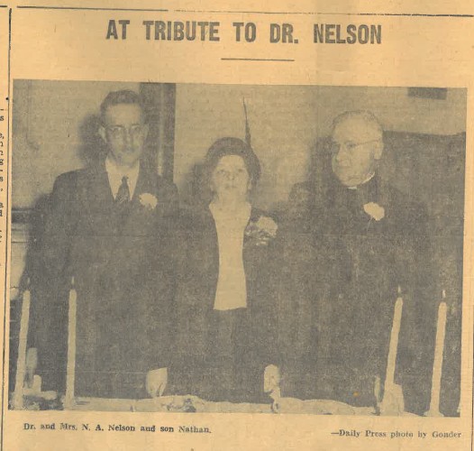 Yellowed newspaper image of older man and woman, with middle-aged man, standing behind a table. Text reads: At Tribute to Dr. Nelson. Dr. and Mrs. NA Nelson and son Nathan.